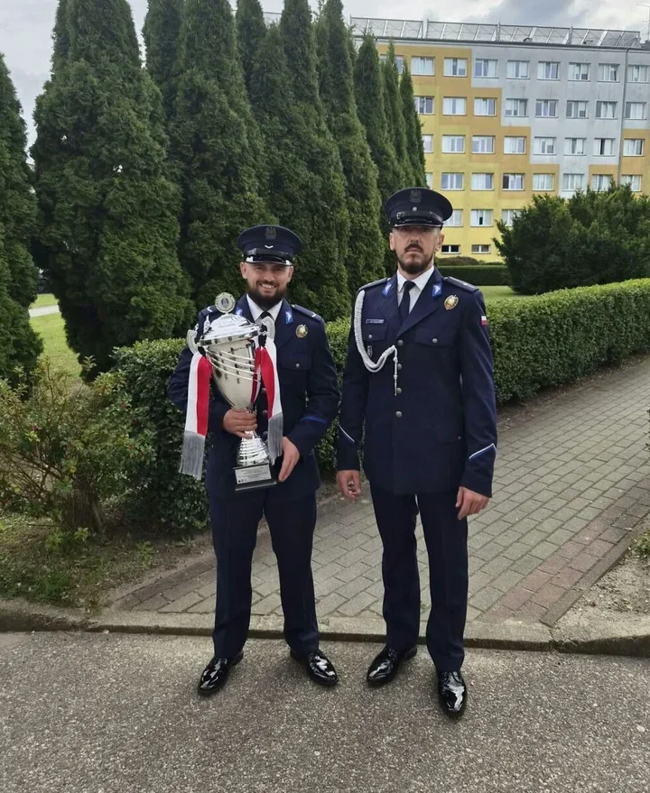 Dwaj mężczyźni w granatowych mundurach z czapkami stoją na chodniku przed zielonymi krzewami i blokiem, jeden z nich trzyma duży srebrny puchar z czerwono-białą