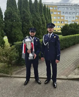 Dwaj mężczyźni w granatowych mundurach z czapkami stoją na chodniku przed zielonymi krzewami i blokiem, jeden z nich trzyma duży srebrny puchar z czerwono-białą