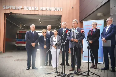 Grupa osób, w tym mężczyzna przemawiający do mikrofonów, stoi przed wejściem do szpitalnego oddziału ratunkowego, w tle widoczna karetka pogotowia.